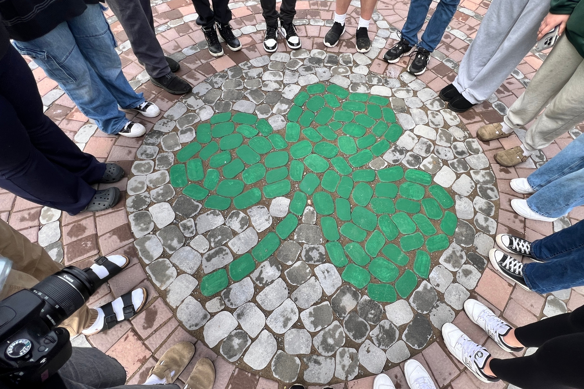 A green shamrock in brick form with the shoes of several people standing around it