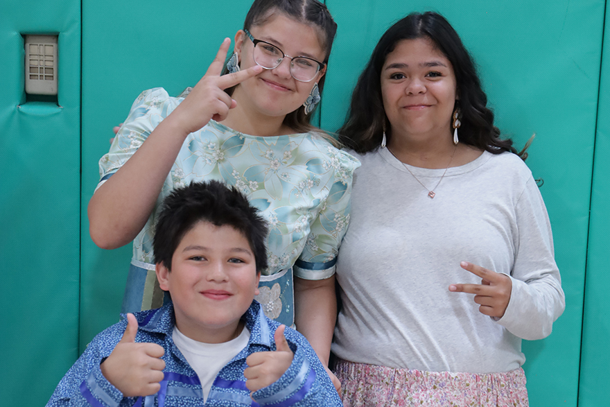 Three students smiling at the camera with peace signs and thumbs up