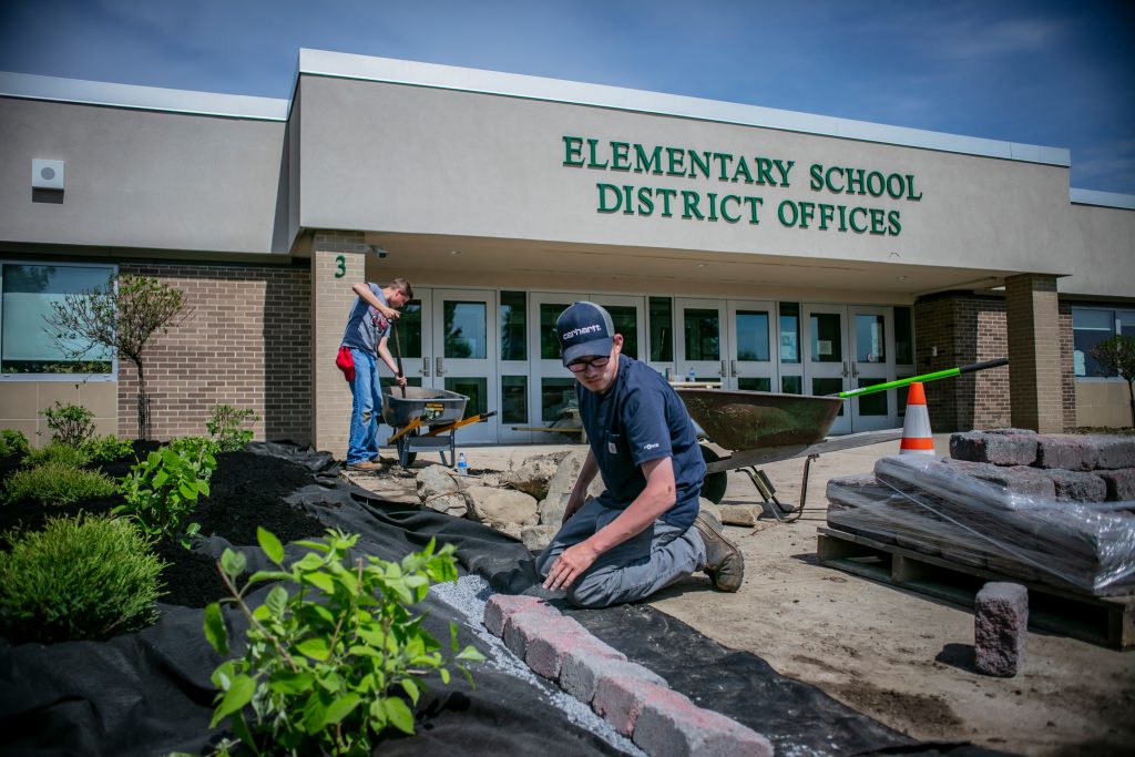Two students work outside on a landscaping project next to a building that says 'Elementary School, District Offices'