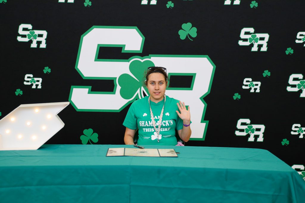 A girl waving while sitting at a table in front of a backdrop with repeated 'SR' letters on it