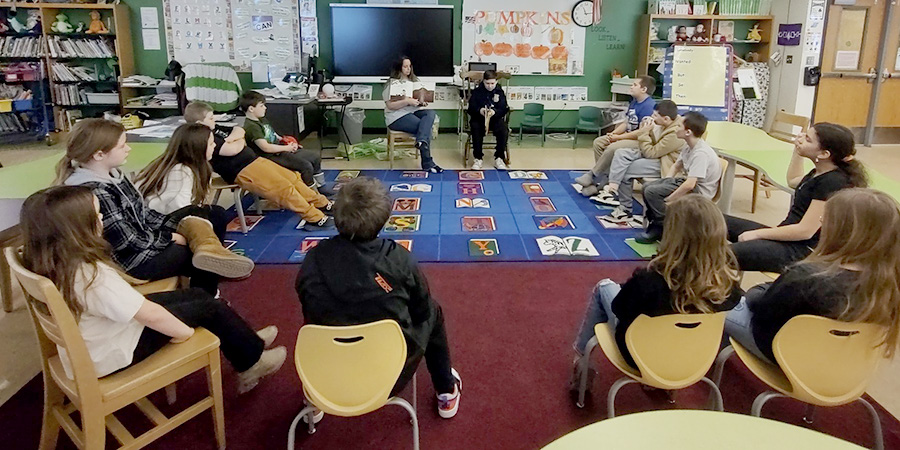 A group of students sitting in chairs in a circle with a teacher reading a book