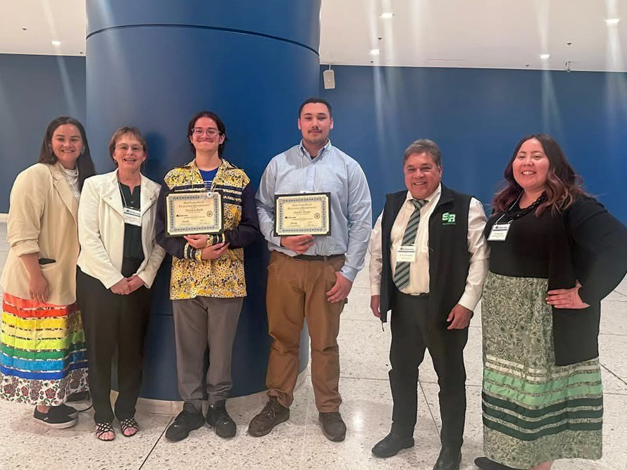 Two students standing while holding certificates posing for a photo alongside four adults
