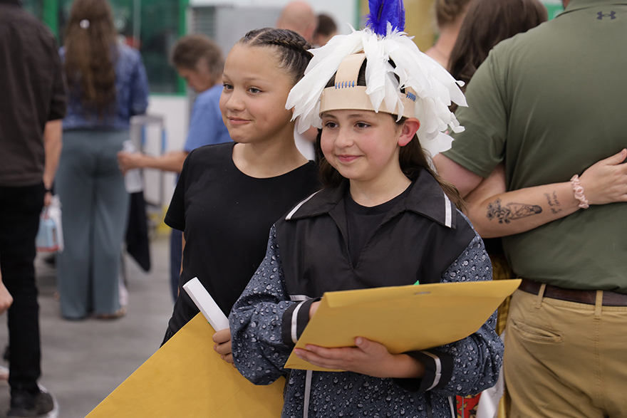 Two students smiling with yellow manilla folders; one with a headdress